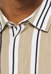 Tans en wit gestreepte shirt met zwarte accenten, voorzien van een geknoopte kraag en zichtbare stikseldetails. De stof lijkt lichtgewicht en textuur te hebben.