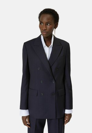 Czarna marynarka dwurzędowa z dopasowanymi spodniami noszona na białej koszuli, stylizowana z długimi cienkimi kolczykami, model lekko patrzy w lewo.