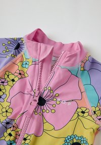 Maillot de bain floral coloré avec un col rose et une fermeture éclair. Le matériau est lisse, avec de grands motifs floraux dans des teintes de rose, jaune et bleu.