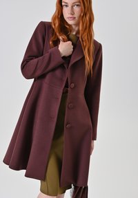 Cappotto bordeaux con un design aderente, tre grandi bottoni e una silhouette svasata. Tessuto liscio, maniche lunghe e rever appuntiti.