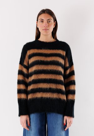 Donna con capelli castano lisci che indossa un maglione peloso a righe nero e marrone e jeans blu, in piedi su sfondo bianco.