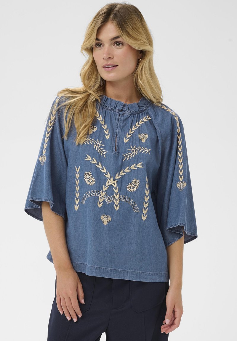 Naine kannab lahtist sinist denimist pluusi, millel on beežid tikandid lillede ja lehtede mustriga, koos tumedate pükste ja ühtlase taustaga.