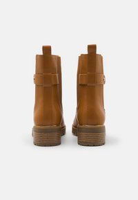 Bottines beiges à talon empilé, présentant une finition en cuir lisse et des sangles décoratives à l'arrière, placées sur un fond uni.
