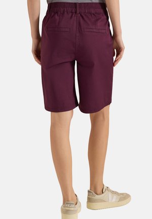 Vista posteriore di una persona che indossa pantaloncini bordeaux fino al ginocchio con cintura elastica e scarpe da ginnastica beige con marchio "VEJA".