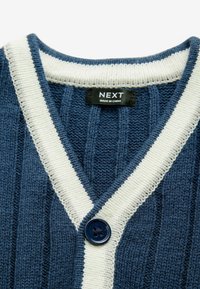 Blauer gestrickter Cardigan mit cremefarbenem Rand, einem einzigen blauen Knopf und einem schwarzen Etikett mit der Aufschrift NEXT made in China am Halsausschnitt.