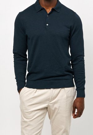 Polo manches longues bleu marine en tissu doux avec un col, trois boutons et un ourlet côtelé, associé à un pantalon de couleur claire.