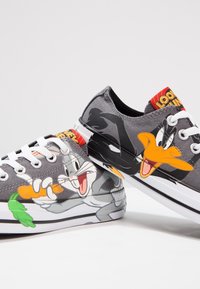 Sneakers in tela grigia con design dei cartoni animati di Bugs Bunny che tiene una carota e Daffy Duck. Lacci bianchi e suole in gomma nera completano il look.