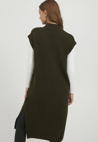 Gilet lavorato a maglia di un verde scuro con una trama a coste, maniche corte e spacchi laterali, indossato sopra un top bianco a maniche lunghe.