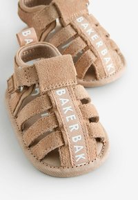 Brune ruskindsandaler med en snørelignende design, tekstilaksenter og hvid branding på siden, med åben tå og polstret sål.