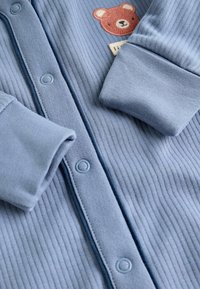 Helles blauer, gerippter Strampler mit Bärenstickerei, Druckknöpfen und umgeschlagenen Ärmeln. Strukturiert Stoff mit glatter Oberfläche und kontrastierender Einfassung.