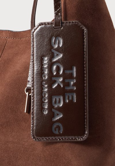 Кафява велурена чанта с тъмнокафява кожена табелка с изписано с ембосинг „"THE SACK BAG MARC JACOBS"“, прикрепена с тънка каишка.