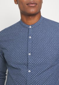 Lähikuva henkilöstä, jolla on päällään sininen nappikauluspaita, jossa on valkoinen geometrinen kuvio ja mandariinikaulus, jolloin näkyy kaula ja ylävartalo.