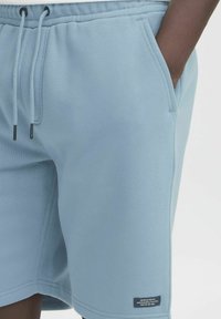 Pantaloni scurți dintr-un material moale de un albastru deschis, cu talie ajustabilă cu șnur, buzunare laterale și o etichetă pe tivul inferior cu detalii textuale.