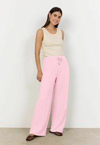 Mujer sonriente, vistiendo una blusa sin mangas beige, pantalones anchos rosa con cordón y zapatos planos negros de malla, parada contra una pared blanca.