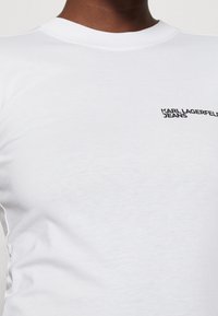 Vitt t-shirt med rund halsringning gjort av mjuk bomull, med en liten svart logotyp "KARL LAGERFELD JEANS" på vänster bröstarea.
