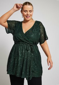 Top avvolgente verde con paillettes, maniche corte, scollatura a V e cintura con fiocco in vita. La texture morbida e la finitura scintillante esaltano il design.