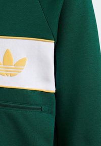 Grön sweatshirts med vita och gula accenter, med en dragkedjeficka och en framträdande logotyp. Tillverkad av texturerat tyg.