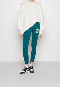 Vit oversized sweatshirt i kombination med teal leggings med Nike-logotyp, kompletterad med svarta och vita sneakers. Släta texturer genom hela outfiten.