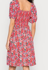 Frau in einem roten Blumenkleid mit kurz gepufften Ärmeln und gerüschtem Rückenteil, vor einem schlichten hellen Hintergrund stehend.