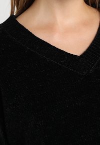 Černý manšestrový top s výstřihem do V, texturovanou látkou a žebrovaným výstřihem, ukazující měkký, plyšový povrch a volný střih.