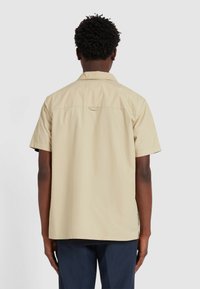 Beige overhemd met korte mouwen en een kraag, voorzien van een enkele plooien aan de achterkant. Het heeft een zachte textuur en een ophanglus bij de nek.