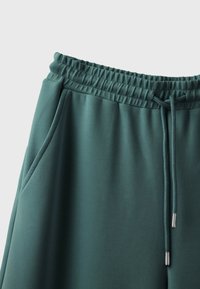 Pantalon de survêtement vert sarcelle avec taille élastique, cordon avec embouts métalliques et poches latérales sur un fond clair uni.