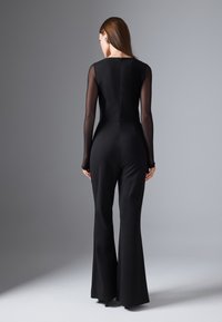 Mono negro con mangas largas de tul, cuerpo ajustado y pantalones acampanados. Presenta un diseño minimalista con cierre de cremallera en la parte posterior.