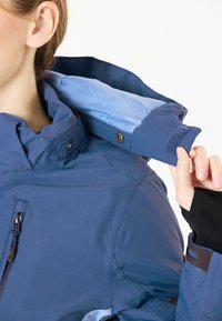 Marinbl�å vattentät jacka med hög krage, blå innerfoder, dragkedjeficka och knäppta ärmdetaljer. Mjuk tygstruktur.