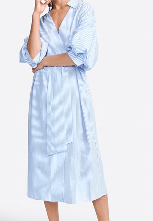 Kobieta ubrana w midi sukienkę koszulową z pionowymi paskami w kolorze jasnoniebieskim i białym, z podwiniętymi rękawami, stojąca na tle jednolitego białego tła.