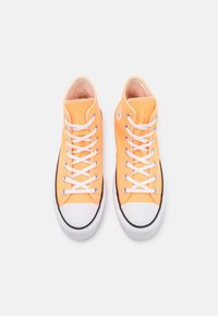 Pomarańczowe trampki w stylu high-top wykonane z płótna, z białymi sznurowadłami, białą gumową osłoną na palce i czarnym paskiem wzdłuż podeszwy. Podeszwa ma teksturowany bieżnik.