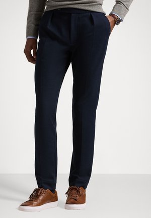 Navy trousers with a pleated design, made of a smooth fabric. Paired with brown leather sneakers and a light grey sweater.
