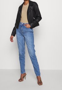 Persona vestida con una chaqueta de cuero negra, una blusa a rayas amarillas y negras, jeans de tiro alto azul y sandalias de tacón verde oliva.