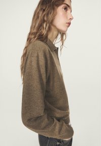 Maglione marrone lavorato a maglia con maniche lunghe e colletto, caratterizzato da una vestibilità comoda e tessuto strutturato. Indossato con jeans scuri casual.