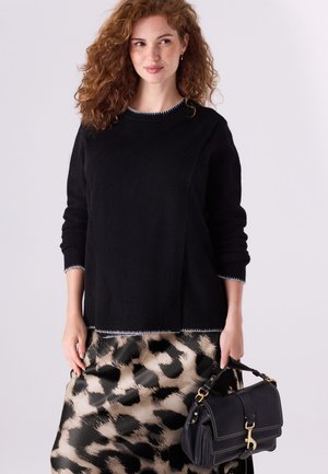 Femme aux cheveux roux bouclés portant un pull noir et une jupe beige-noir à motif animalier, tenant un sac à main noir sur fond uni.