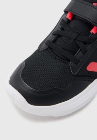 Czarny sportowy but z siateczkową cholewką i syntetycznymi dodatkami. Z czerwonymi akcentami, zapięciem na rzep i białą gumową podeszwą. Strukturalna powierzchnia.