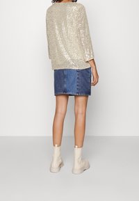 Top beige con paillettes e maniche a tre quarti, abbinato a una gonna di jeans. Stivaletti bianchi alla caviglia completano l'outfit. Sfondo neutro.