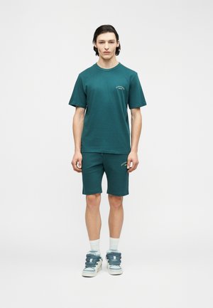 Νέος άνδρας που στέκεται, φοράει μπλουζάκι κοντό μανίκι σε τυρκουάζ χρώμα και αντίστοιχο σορτς, λευκές κάλτσες και χοντρά αθλητικά παπούτσια με μπλε κορδόνια, σε λευκό απλό φόντο.