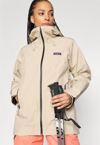 Bež veekindel jope kõrge kraega, musta tõmblukuga, reguleeritava kapuutsi ja küljetaskutega, kantud matkakeppidega.