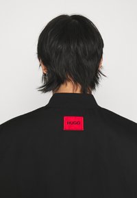 Giacca nera con colletto alto e un evidente logo rettangolare rosso etichettato "HUGO" sul retro. Texture liscia e design elegante.