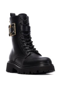 Botín de cuero negro hasta el tobillo con cordones en la parte frontal, suela gruesa y un prominente hebilla decorativa en negro y dorado en el lateral.