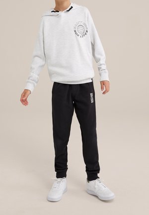Kind trägt weißen Hoodie mit dem Schriftzug „Salty Dog“ und schwarze Jogginghose mit weißen Sneakers, steht auf beige-farbenem Hintergrund.