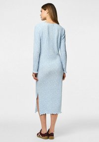 Vestido de manga larga en tejido de canalé azul claro con un patrón floral, abertura lateral y dobladillo con bordes en forma de ondas, combinado con zapatillas burdeos.