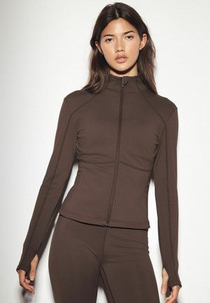 Frau trägt eine eng anliegende dunkelbraune Jacke mit Reißverschluss und passende Hose, steht vor einem schlichten hellen Hintergrund.