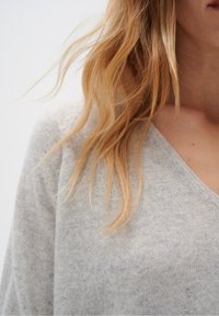 Cheveux blonds tombant sur un pull gris clair à col en V, sur une personne dont on aperçoit partiellement le cou et le bas du visage.