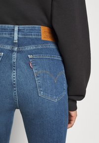Jean en denim bleu à taille haute, avec des poches arrière, un patch en cuir marron clair et des coutures distinctives sur les poches.