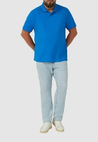 Mężczyzna stoi z rękami za plecami, ubrany w jasnoniebieską koszulę polo, jasnoniebieskie dżinsy i białe sneakersy na jednolitym tle.