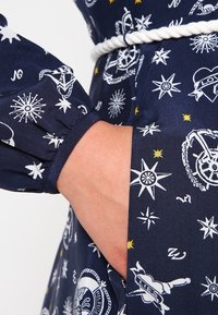 Dunkelblaues Kleid mit weißen nautisch inspirierten Mustern, gerafften Ärmel-Details und einem Taillengürtel. Glattes Gewebe mit einer weichen Textur.