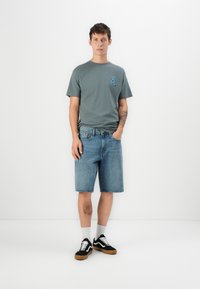 Ung mand står op ad en hvid væg iført en grå grafisk t-shirt, denimshorts, hvide sokker og sorte sneakers med gummisåler.