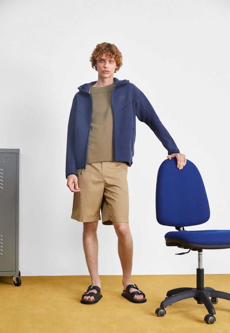 Sweat à fermeture éclair navy par-dessus une chemise vert olive, un short beige et des sandales noires. Une chaise de bureau bleue est positionnée à côté de l'individu.