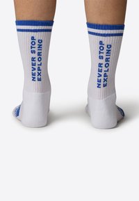 Białe skarpetki z niebieskimi paskami na górze, z napisem "NIGDY NIE PRZESTAWAJ ODKRYWAĆ" wplecionym na niebiesko po bokach. Miękka, prążkowana faktura.